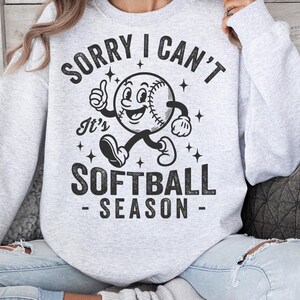 Op de afbeelding: Lichtgrijs sweatshirt met de tekst "SORRY I CAN'T It's SOFTBALL SEASON" in het zwart. Het ontwerp toont een cartoon honkbalpersonage dat een duim omhoog geeft. Het sweatshirt is een casual, comfortabele stijl.