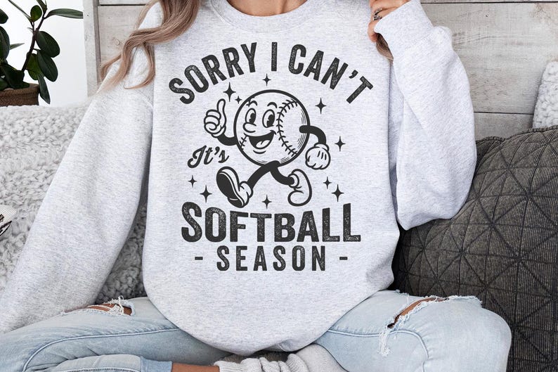 Op de afbeelding: Lichtgrijs sweatshirt met de tekst "SORRY I CAN'T It's SOFTBALL SEASON" in het zwart. Het ontwerp toont een cartoon honkbalpersonage dat een duim omhoog geeft. Het sweatshirt is een casual, comfortabele stijl.
