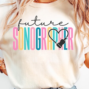 Puede incluir: Camiseta color crema con las palabras "future SONOGRAPHER" en un diseño colorido. La palabra "future" está en una fuente manuscrita, y "SONOGRAPHER" está en una fuente de bloque en negrita y multicolor. Una imagen de sonograma en forma de corazón negro está en el centro.