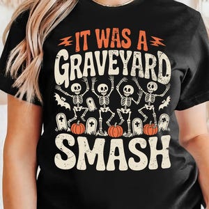 May include: Black t-shirt with a Halloween-themed graphic. The design features the text "IT WAS A GRAVEYARD SMASH" in orange and white, along with illustrations of skeletons, pumpkins, and tombstones. A festive and spooky design.