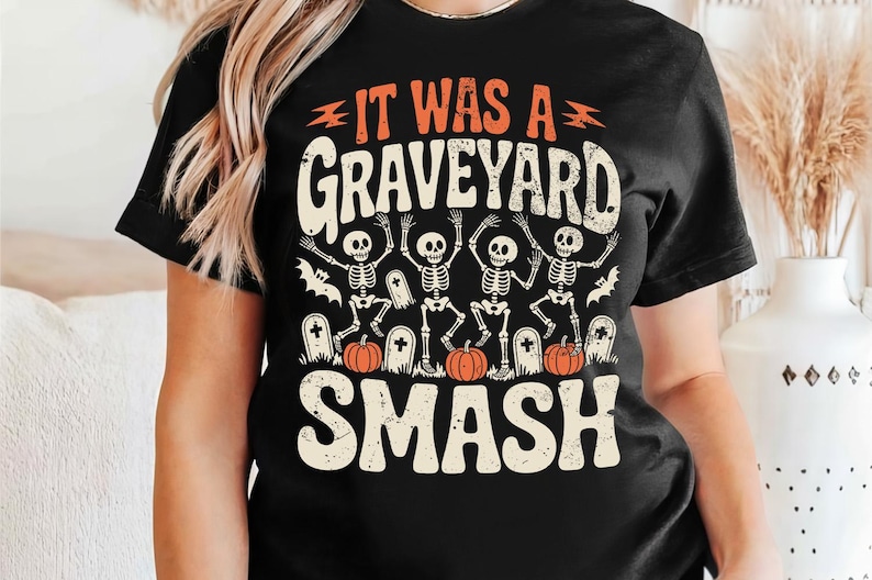 May include: Black t-shirt with a Halloween-themed graphic. The design features the text "IT WAS A GRAVEYARD SMASH" in orange and white, along with illustrations of skeletons, pumpkins, and tombstones. A festive and spooky design.