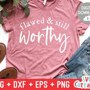 Puede incluir: Una camiseta rosa con el texto "flawed & still worthy" en letras blancas. La camiseta está sobre una superficie blanca con un par de sandalias marrones y unos vaqueros azules.