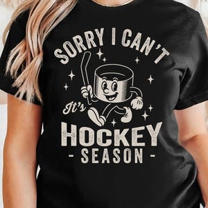 May include: Black t-shirt with the text "SORRY I CAN'T It's HOCKEY SEASON". The graphic features a cartoon hockey puck character holding a hockey stick and running. The design includes stars.