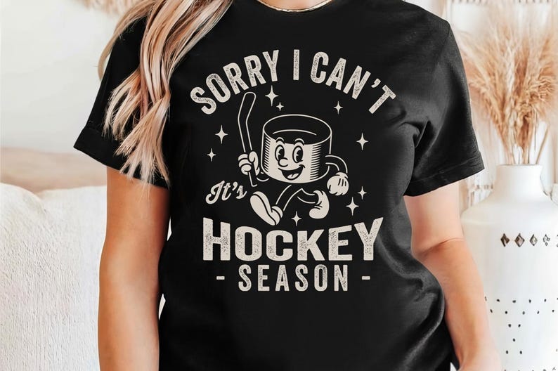 May include: Black t-shirt with the text "SORRY I CAN'T It's HOCKEY SEASON". The graphic features a cartoon hockey puck character holding a hockey stick and running. The design includes stars.