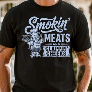 May include: Black t-shirt with a white graphic. The graphic features the text "Smokin' MEATS and CLAPPIN' CHEEKS" with a cartoon pig chef holding a tray of ribs. The pig is wearing a chef's hat and apron.