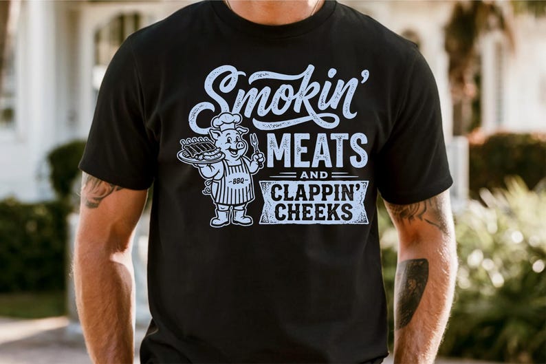 May include: Black t-shirt with a white graphic. The graphic features the text "Smokin' MEATS and CLAPPIN' CHEEKS" with a cartoon pig chef holding a tray of ribs. The pig is wearing a chef's hat and apron.