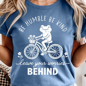 May include: A blue t-shirt featuring a white illustration of a frog on a bicycle carrying flowers. The shirt has the text "BE HUMBLE BE KIND" and "leave your worries BEHIND" printed on it.
