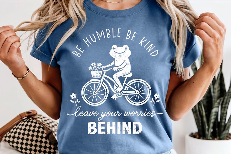 May include: A blue t-shirt featuring a white illustration of a frog on a bicycle carrying flowers. The shirt has the text "BE HUMBLE BE KIND" and "leave your worries BEHIND" printed on it.