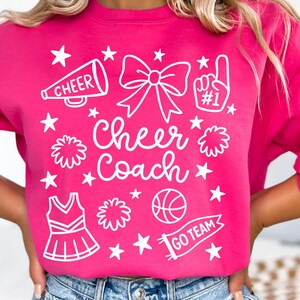May include: A bright pink sweatshirt with white graphics. The design includes a megaphone, bow, pom-pom, cheerleader uniform, basketball, and the words "Cheer Coach" and "Go Team". Stars are scattered throughout the design.