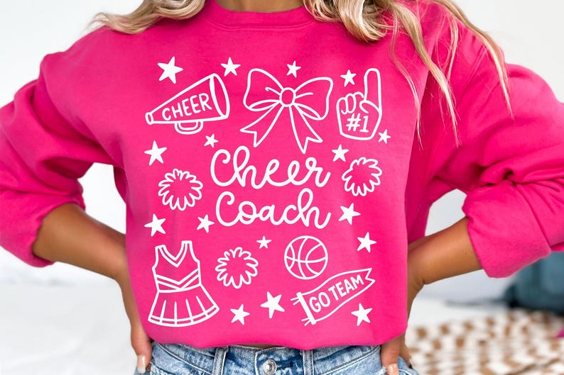 May include: A bright pink sweatshirt with white graphics. The design includes a megaphone, bow, pom-pom, cheerleader uniform, basketball, and the words "Cheer Coach" and "Go Team". Stars are scattered throughout the design.