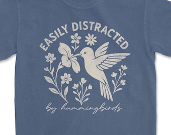 Può includere: Maglietta blu con grafica bianca di un colibrì, fiori e la scritta "EASILY DISTRACTED by hummingbirds". Il disegno è centrato sulla maglietta.