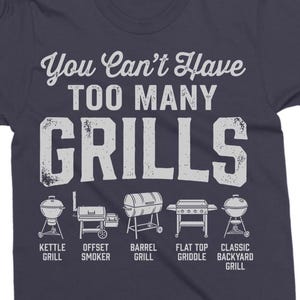 May include: Navy blue t-shirt with the text "You Can't Have TOO MANY GRILLS" in white. Below the text are illustrations of different grill types: kettle, offset smoker, barrel, flat top griddle, and classic backyard grill.