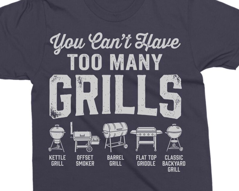 May include: Navy blue t-shirt with the text "You Can't Have TOO MANY GRILLS" in white. Below the text are illustrations of different grill types: kettle, offset smoker, barrel, flat top griddle, and classic backyard grill.