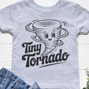 Peut inclure: Un t-shirt gris clair pour enfant avec un dessin de tornade noire et les mots "Tiny Tornado". Le t-shirt est pr&eacute;sent&eacute; avec un jean bleu sur une surface en bois blanc.