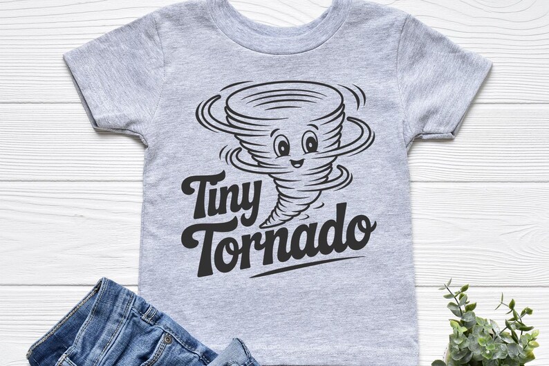 Peut inclure: Un t-shirt gris clair pour enfant avec un dessin de tornade noire et les mots "Tiny Tornado". Le t-shirt est pr&eacute;sent&eacute; avec un jean bleu sur une surface en bois blanc.