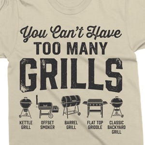 May include: Tan t-shirt with the text "You Can't Have TOO MANY GRILLS" in black. Below the text are illustrations of different grill types, including kettle, offset smoker, barrel, flat top griddle, and classic backyard grills.