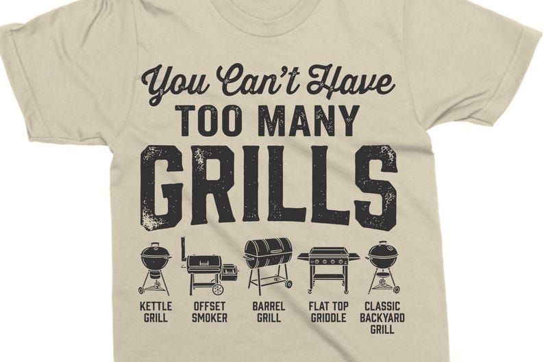 May include: Tan t-shirt with the text "You Can't Have TOO MANY GRILLS" in black. Below the text are illustrations of different grill types, including kettle, offset smoker, barrel, flat top griddle, and classic backyard grills.