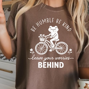 May include: A brown t-shirt featuring a white graphic of a frog on a bicycle, a basket of flowers, and the phrases "BE HUMBLE BE KIND" and "leave your worries BEHIND". The shirt has a relaxed fit and a neutral colour.