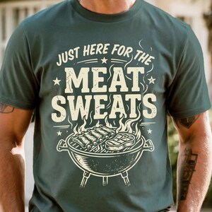 May include: Dark green t-shirt with the text "JUST HERE FOR THE MEAT SWEATS" in a distressed, vintage style. The design includes a grill with ribs and steak. A casual tee for grilling enthusiasts.