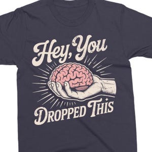 May include: Navy blue t-shirt with a graphic design. The design features a pink brain held in a hand, with the text "Hey, You Dropped This" in a white, vintage-style font. The shirt is made of a soft material.
