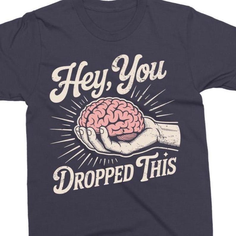 May include: Navy blue t-shirt with a graphic design. The design features a pink brain held in a hand, with the text "Hey, You Dropped This" in a white, vintage-style font. The shirt is made of a soft material.