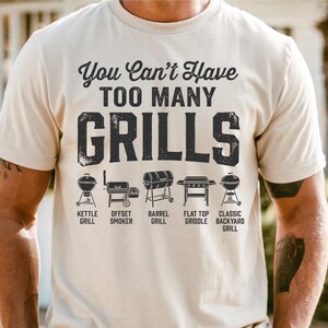 May include: A beige t-shirt with the text "You Can't Have TOO MANY GRILLS" in black. Below the text are illustrations of different grill types: kettle, offset smoker, barrel, flat top griddle, and classic backyard grill.