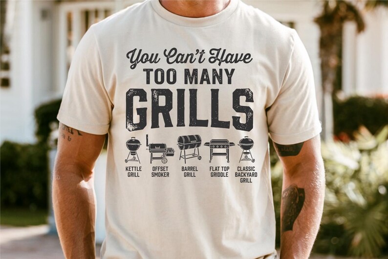 May include: A beige t-shirt with the text "You Can't Have TOO MANY GRILLS" in black. Below the text are illustrations of different grill types: kettle, offset smoker, barrel, flat top griddle, and classic backyard grill.