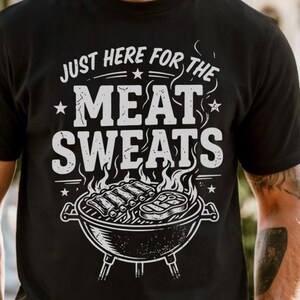 May include: Black t-shirt with a white graphic that reads "JUST HERE FOR THE MEAT SWEATS." The design includes a grill with ribs and steak, flames, and stars. A casual tee for grilling enthusiasts.