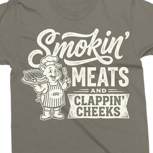 May include: Gray t-shirt with a graphic featuring a cartoon pig chef holding a plate of ribs and a fork. The text reads "Smokin' MEATS and CLAPPIN' CHEEKS" in a vintage style.