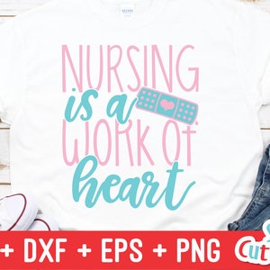 Pu&ograve; includere: Una maglietta bianca con il testo "NURSING is a WORK of heart" in lettere rosa e blu. Un cerotto rosa e blu con un cuore &egrave; accanto al testo. La maglietta &egrave; su una superficie di legno con un paio di scarpe da ginnastica bianche e jeans blu.