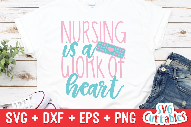 Pu&ograve; includere: Una maglietta bianca con il testo "NURSING is a WORK of heart" in lettere rosa e blu. Un cerotto rosa e blu con un cuore &egrave; accanto al testo. La maglietta &egrave; su una superficie di legno con un paio di scarpe da ginnastica bianche e jeans blu.