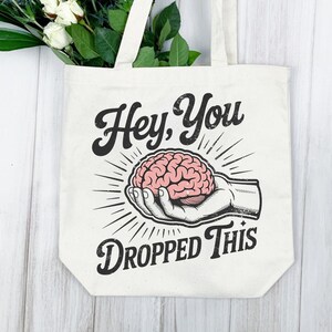 May include: Cream-colored tote bag with the text "Hey, You Dropped This" in a vintage style. The graphic features a hand holding a pink brain, with radiating lines. White flowers and greenery are visible.