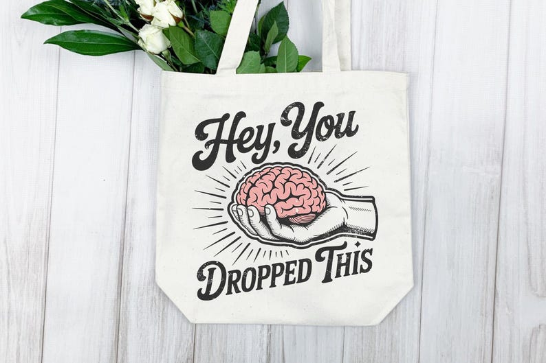 May include: Cream-colored tote bag with the text "Hey, You Dropped This" in a vintage style. The graphic features a hand holding a pink brain, with radiating lines. White flowers and greenery are visible.