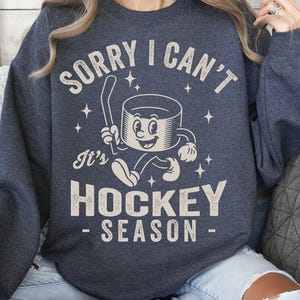 May include: Dark grey sweatshirt featuring the text "SORRY I CAN'T" above a cartoon hockey puck character holding a hockey stick. Below the character is the text "It's HOCKEY SEASON". The design is in a vintage style.