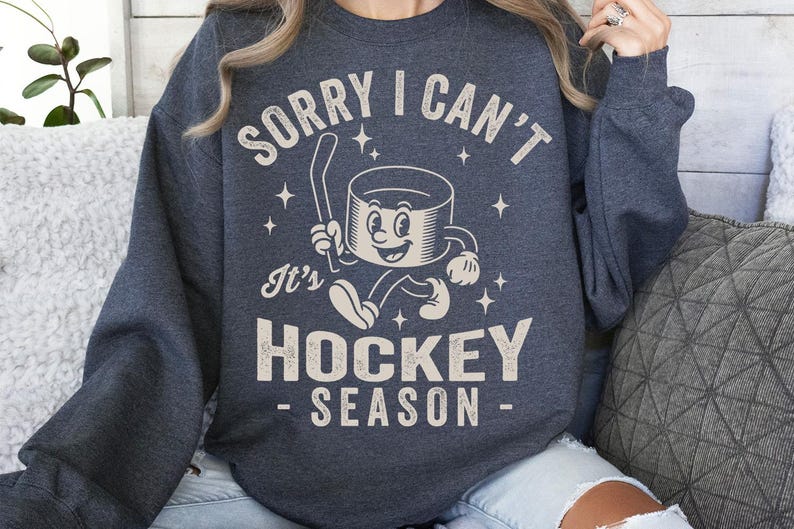 May include: Dark grey sweatshirt featuring the text "SORRY I CAN'T" above a cartoon hockey puck character holding a hockey stick. Below the character is the text "It's HOCKEY SEASON". The design is in a vintage style.
