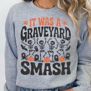 May include: A light gray sweatshirt with a Halloween-themed graphic. The graphic features the text "IT WAS A GRAVEYARD SMASH" in orange and black, along with dancing skeletons, pumpkins, and bats.
