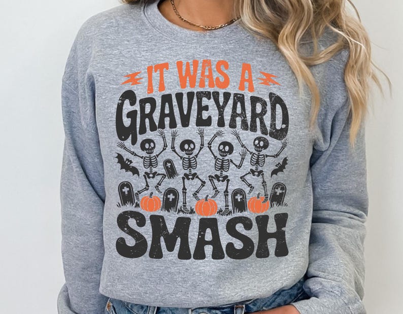 May include: A light gray sweatshirt with a Halloween-themed graphic. The graphic features the text "IT WAS A GRAVEYARD SMASH" in orange and black, along with dancing skeletons, pumpkins, and bats.