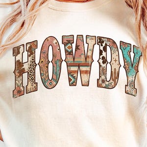 May include: A cream-coloured sweatshirt featuring the word "HOWDY" in a decorative, Western-themed font. The letters are filled with various patterns, including floral, cowhide, and geometric designs. The overall aesthetic is vintage and country-inspired.
