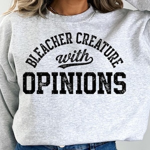 May include: A light gray sweatshirt with the text "BLEACHER CREATURE with OPINIONS" in a black, distressed font. The text is arched across the top and bottom of the design. The sweatshirt has a crew neck.