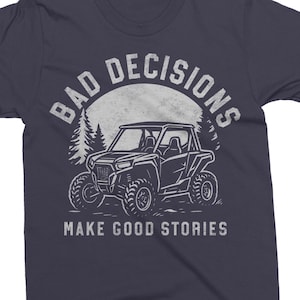 May include: Navy blue t-shirt with a white graphic. The graphic features a side-view illustration of an off-road vehicle, a forest silhouette, and the text "BAD DECISIONS" above and "MAKE GOOD STORIES" below.