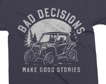 May include: Navy blue t-shirt with a white graphic. The graphic features a side-view illustration of an off-road vehicle, a forest silhouette, and the text "BAD DECISIONS" above and "MAKE GOOD STORIES" below.