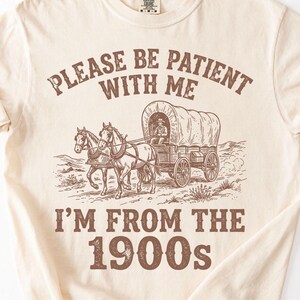 May include: Cream-colored t-shirt with a brown graphic of a covered wagon pulled by two horses. The text reads "PLEASE BE PATIENT WITH ME" above the wagon and "I'M FROM THE 1900s" below. A vintage-style design.