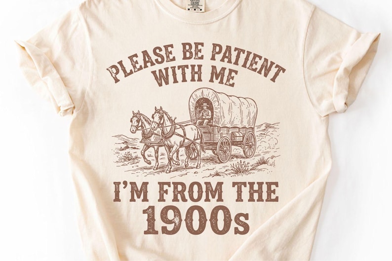 May include: Cream-colored t-shirt with a brown graphic of a covered wagon pulled by two horses. The text reads "PLEASE BE PATIENT WITH ME" above the wagon and "I'M FROM THE 1900s" below. A vintage-style design.