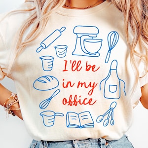 Puede incluir: Camiseta color crema con dibujos lineales azules de utensilios de cocina y una hogaza de pan. El texto "I'll be in my office" está en rojo y azul. La camiseta es de estilo casual y cómodo.
