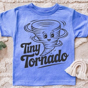 Peut inclure: T-shirt bleu clair avec un dessin anim&eacute; de tornade souriante et l'inscription "Tiny Tornado" en noir. Le t-shirt est en tissu doux.