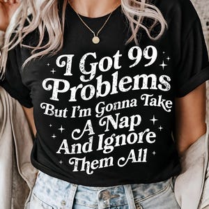 May include: Black t-shirt with the white text "I Got 99 Problems But I'm Gonna Take A Nap And Ignore Them All." The shirt has a crew neck and short sleeves. The text is in a whimsical font with star accents.