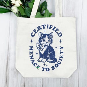 May include: Cream-colored tote bag with a navy blue graphic of a cat wearing a bow tie, surrounded by the words "CERTIFIED MENACE TO SOCIETY." The bag has white handles and is filled with greenery and white flowers.