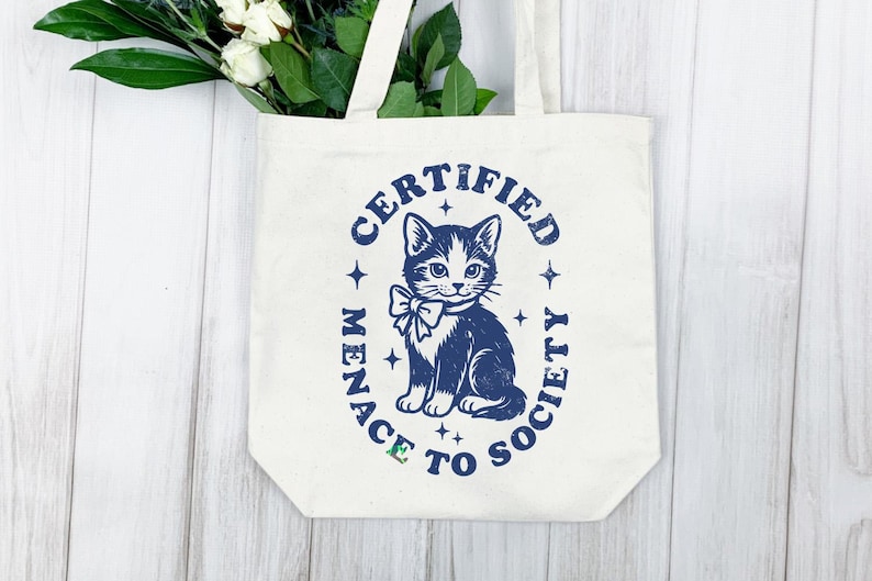 May include: Cream-colored tote bag with a navy blue graphic of a cat wearing a bow tie, surrounded by the words "CERTIFIED MENACE TO SOCIETY." The bag has white handles and is filled with greenery and white flowers.