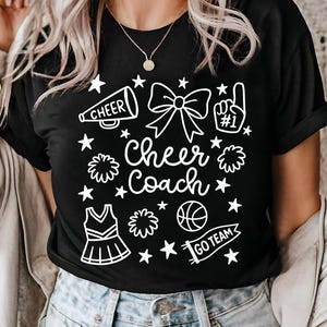 May include: Black t-shirt with a white graphic design featuring cheerleading elements. The design includes a megaphone, bow, pom-poms, a basketball, a cheerleader uniform, stars, and the words "Cheer Coach" and "Go Team".