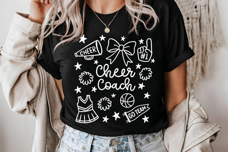 May include: Black t-shirt with a white graphic design featuring cheerleading elements. The design includes a megaphone, bow, pom-poms, a basketball, a cheerleader uniform, stars, and the words "Cheer Coach" and "Go Team".